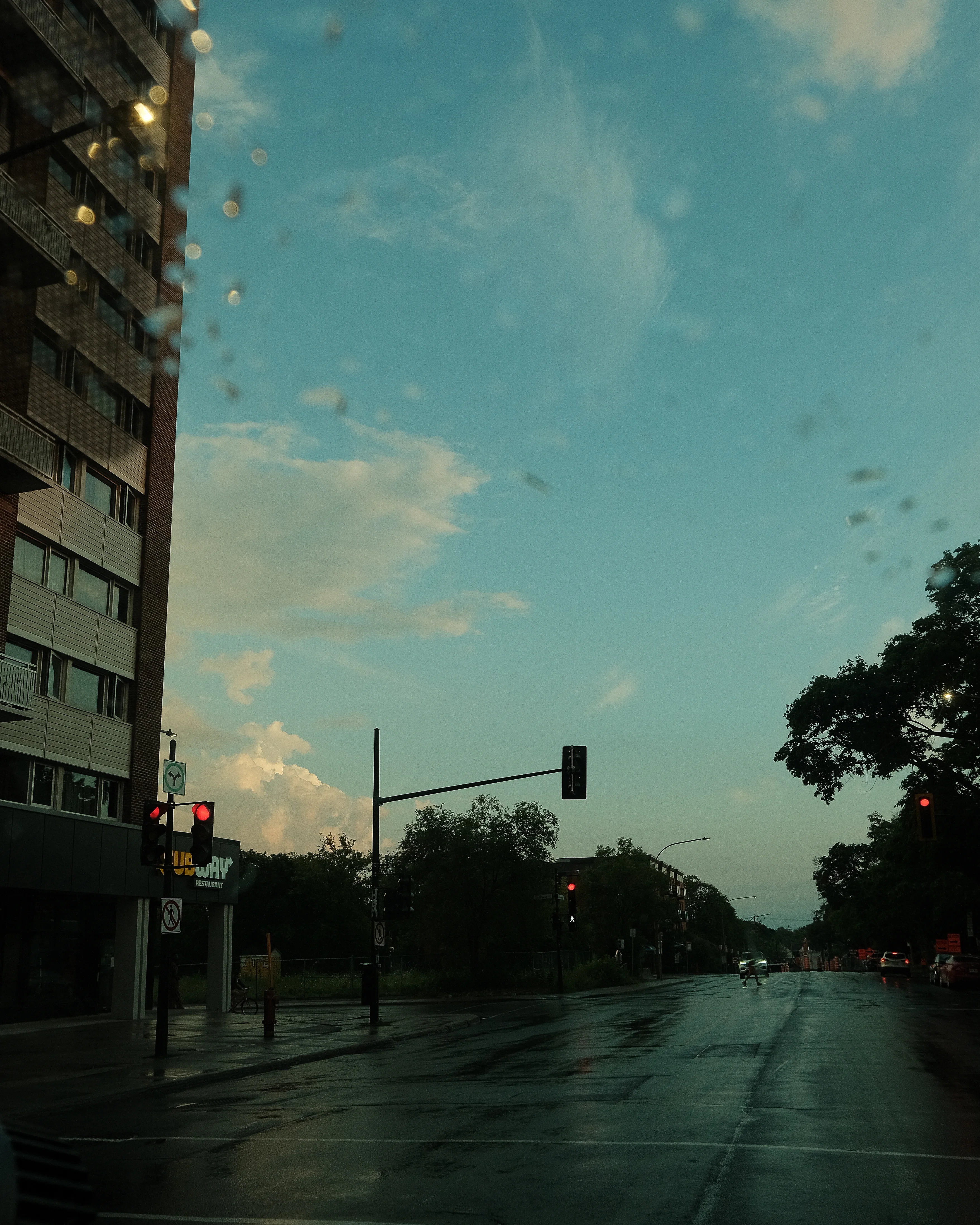 A photo of a busy street at sunset. The sky is a mix of blue and orange, and there are rain-soaked roads and traffic lights. Buildings and trees are visible on either side of the street.